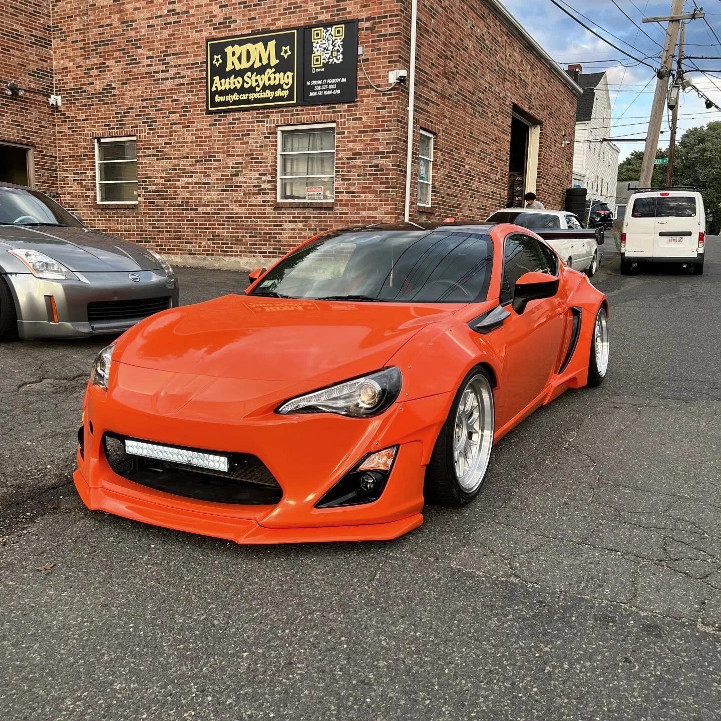 Orange FRS with a professional vinyl vehicle wrap parked on asphalt with brick building and other vehicles in the background.