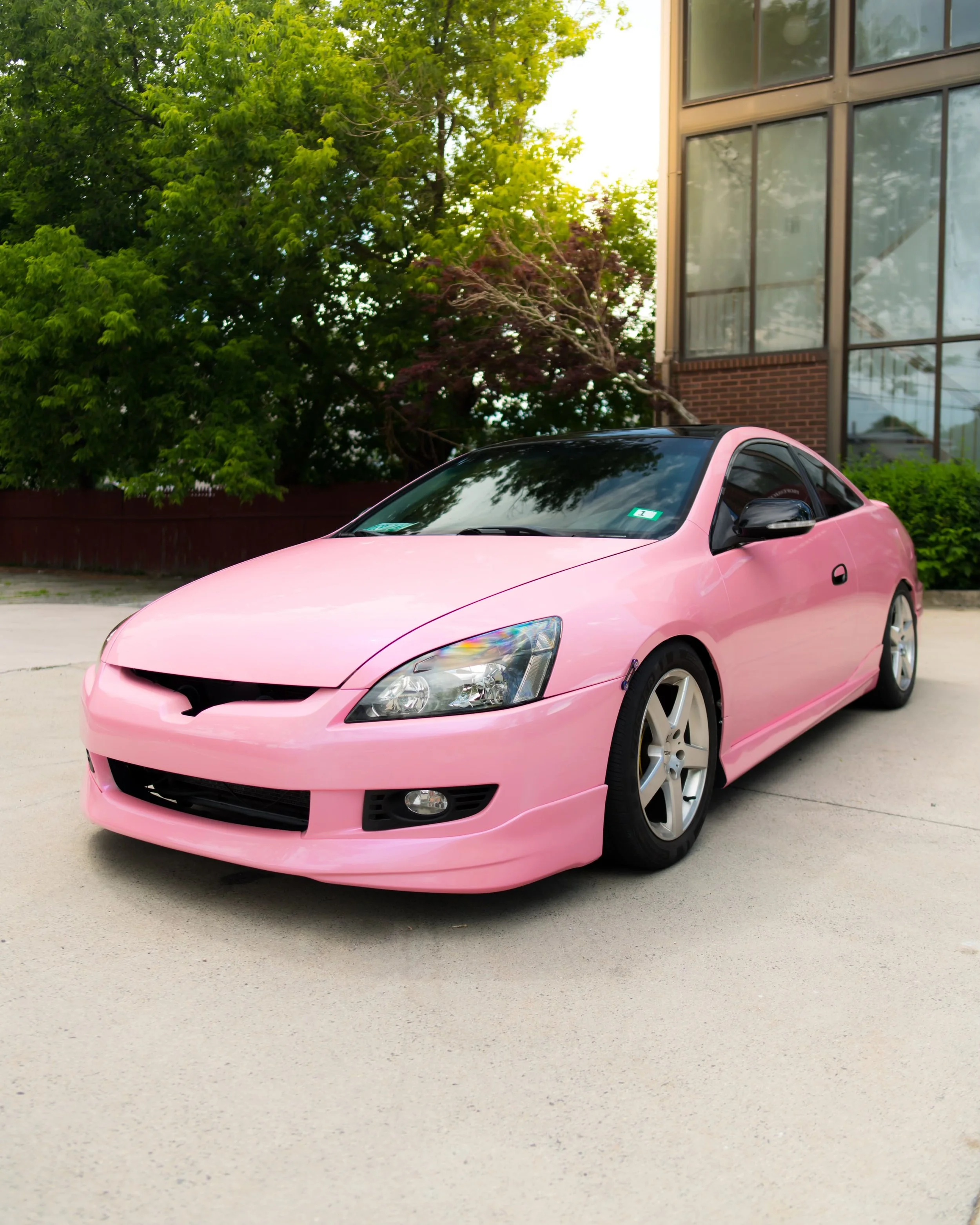 A pink sports car parked on a concrete surface next to a green bush and trees, with a modern building in the background.