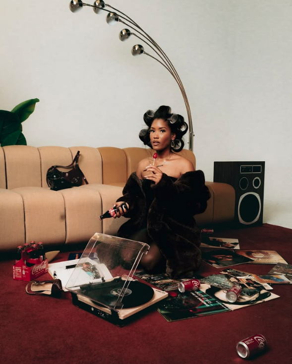 A woman with hair rollers in her hair is sitting on a carpeted floor in a living room, surrounded by magazines and soda cans, holding a bottle and a remote, with a beige couch and a floor lamp in the background.