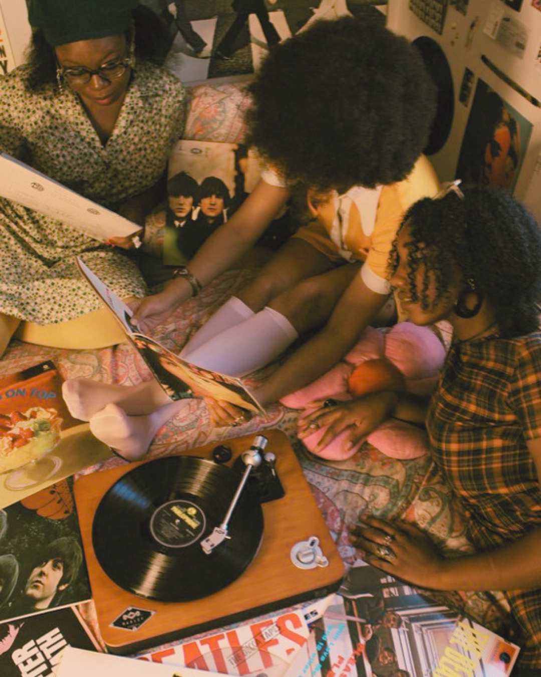 Three people, likely a family, are gathered around a vintage record player on a bed, looking at and handling vinyl records. The scene has a nostalgic, retro vibe with posters on the wall and magazines scattered around.