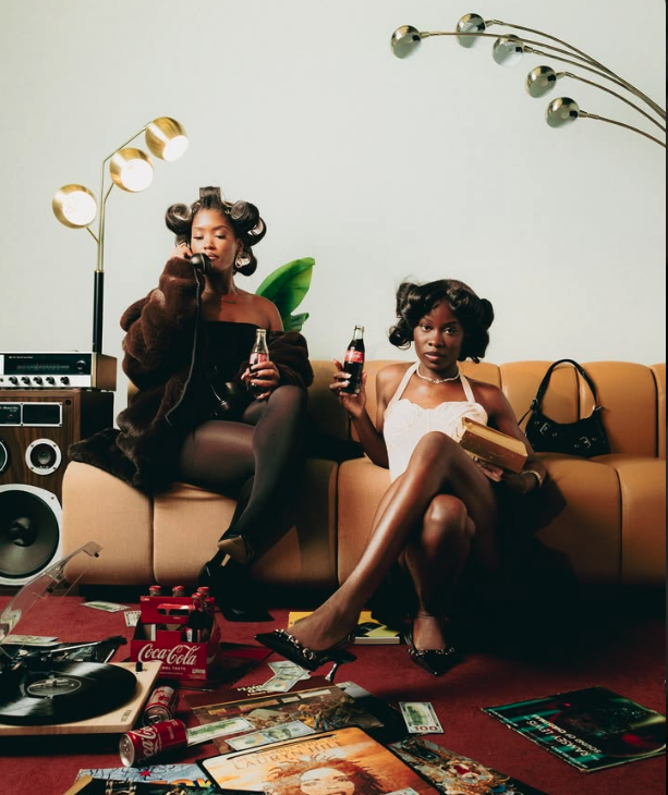 Two women sitting on a beige couch in a room with vintage decor, holding bottles of soda. One woman is wearing a black dress and fur coat, the other is in a white dress with high heels. There are magazines, a record player, and scattered dollar bills
