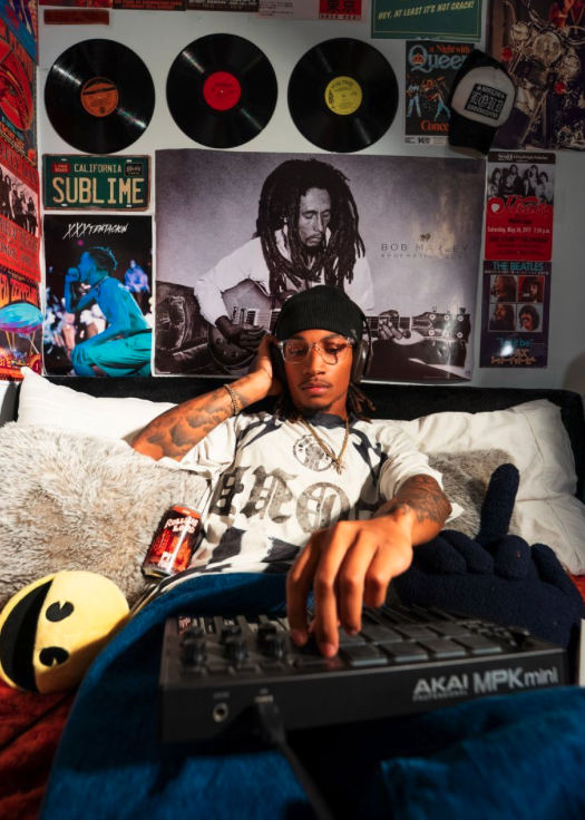 Young man with dreadlocks, wearing glasses, a black cap, and a white T-shirt, listening to headphones, lying on a bed with various posters of music artists and vinyl records on the wall behind him, and using music production equipment.