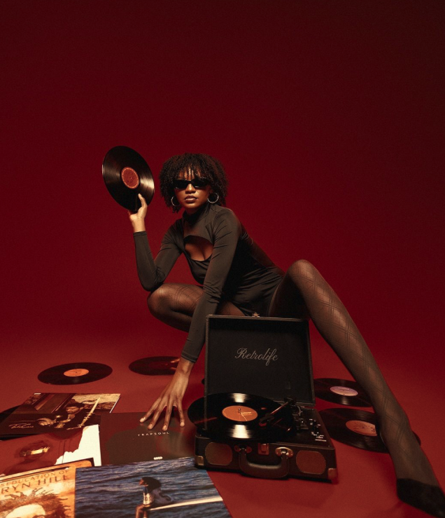 A woman with curly hair, wearing sunglasses, black outfit, fishnet tights, and hoop earrings, sitting on the floor surrounded by vinyl records and a vintage record player, with a solid red background.
