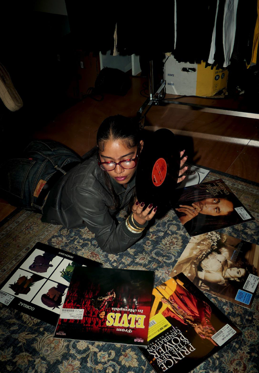 Person lying on a rug, flipping through vinyl records surrounded by more records with album covers visible.