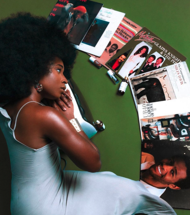 A woman with curly hair lying on her side on a green table, surrounded by vinyl records, old magazines, and perfume bottles.
