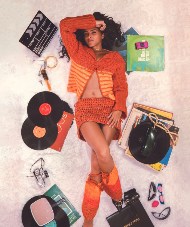 A young woman lying on a soft white surface surrounded by vintage records, a movie clapperboard, a camera, headphones, sunglasses, and makeup. She is wearing an orange knitted cardigan, skirt, and leg warmers.