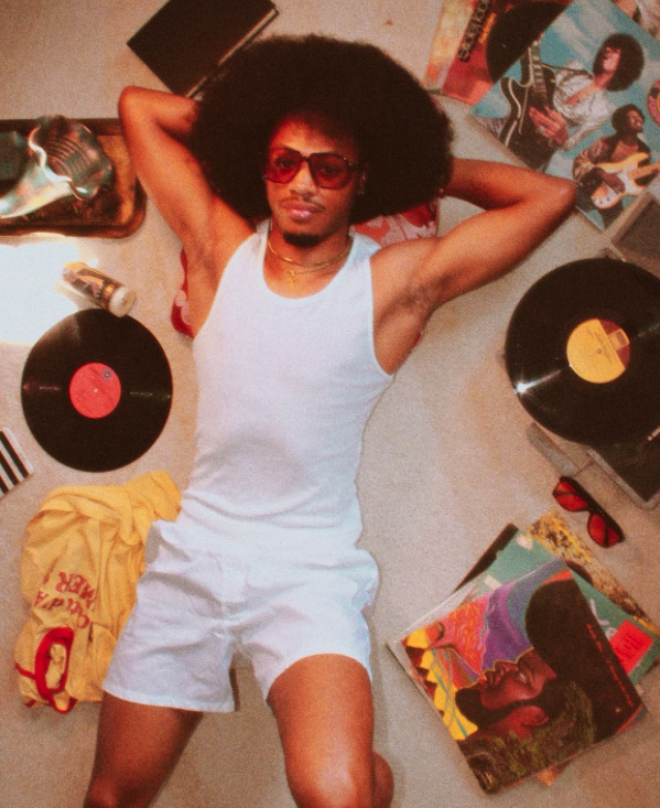 A man with curly afro hair and sunglasses, lying on the floor, wearing a white sleeveless shirt and shorts, surrounded by vinyl records, colorful posters, sunglasses, and a yellow shirt.