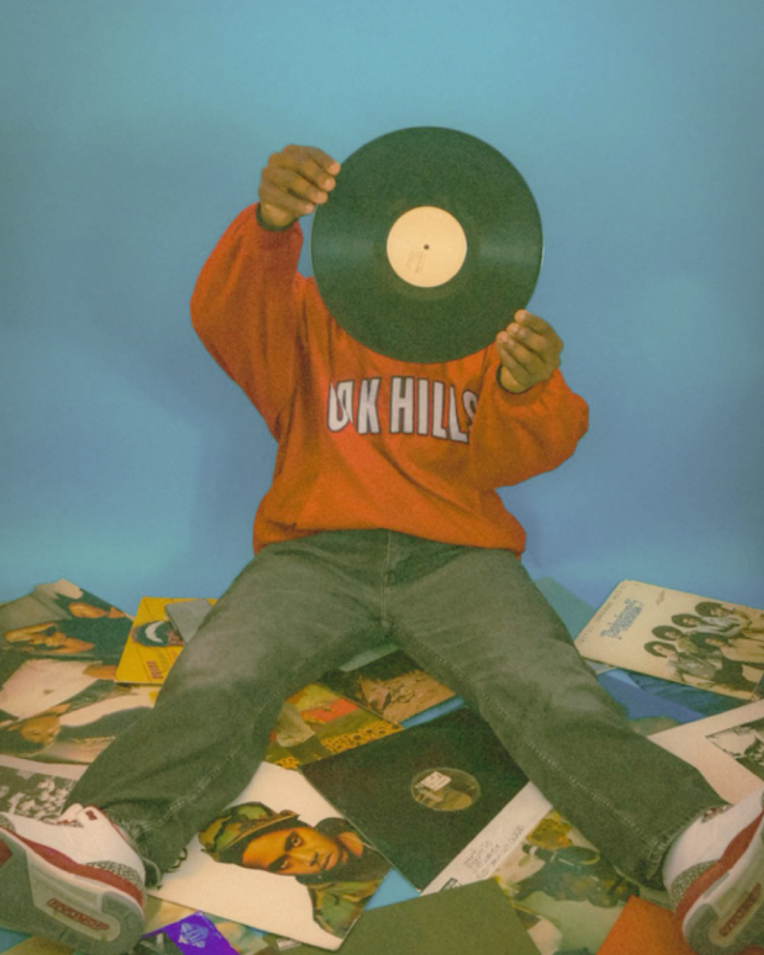 Person sitting with legs spread apart on floor covered with records, holding a vinyl record in front of their face, wearing an orange sweatshirt with 'OK HILLS' written on it, against a blue wall.