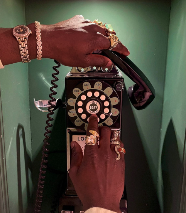 Close-up of a person's hand with multiple rings, pressing a rotary dial on a vintage black payphone with a coiled cord, inside a green phone booth.