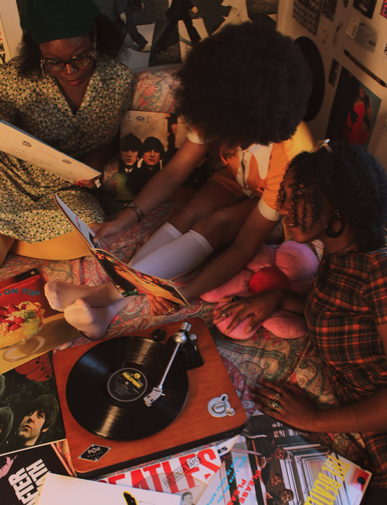 Three women with curly hair and glasses sitting on a bed, looking at vinyl record covers, with a turntable on a nearby table, surrounded by vinyl records and posters.