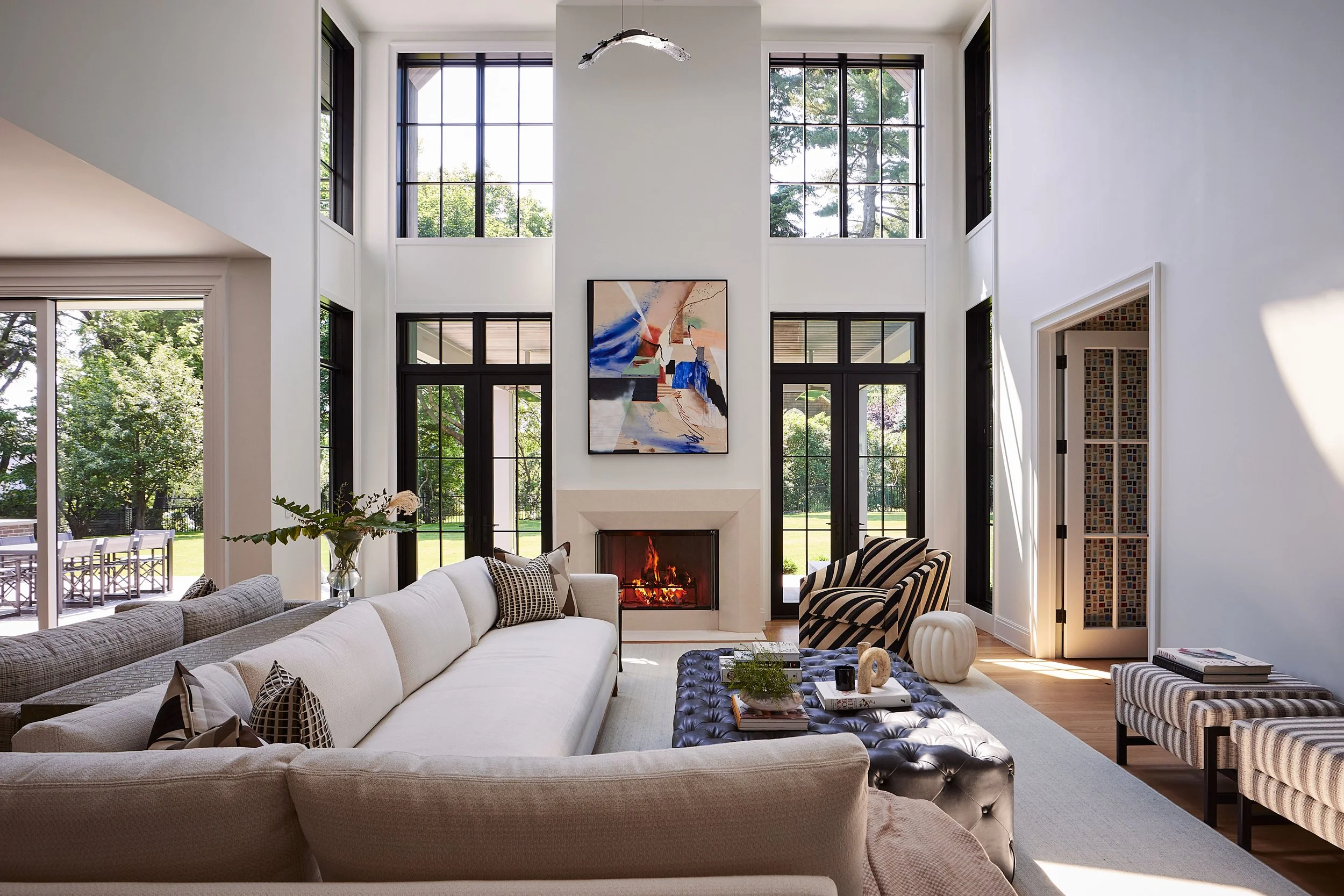 Bright living room with white walls, large black-framed windows, and a fireplace. Contains a large white sofa, a black tufted ottoman, a striped armchair, and a side table. Decorations include a large abstract painting, a dark striped chair, and a va