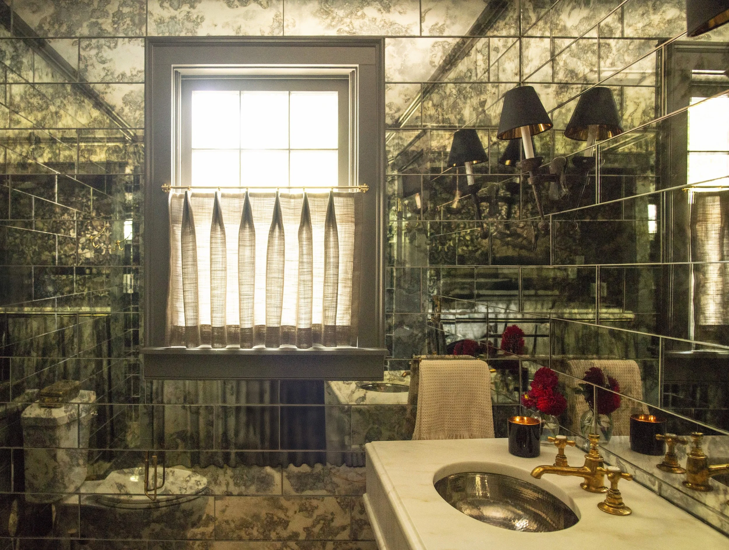 A vintage-style bathroom with a window covered by sheer curtains, reflecting mirrors on the walls, a marble sink with gold fixtures, and black lamps attached to the mirror. There are red flowers on the sink, and marble statues on the ledge.
