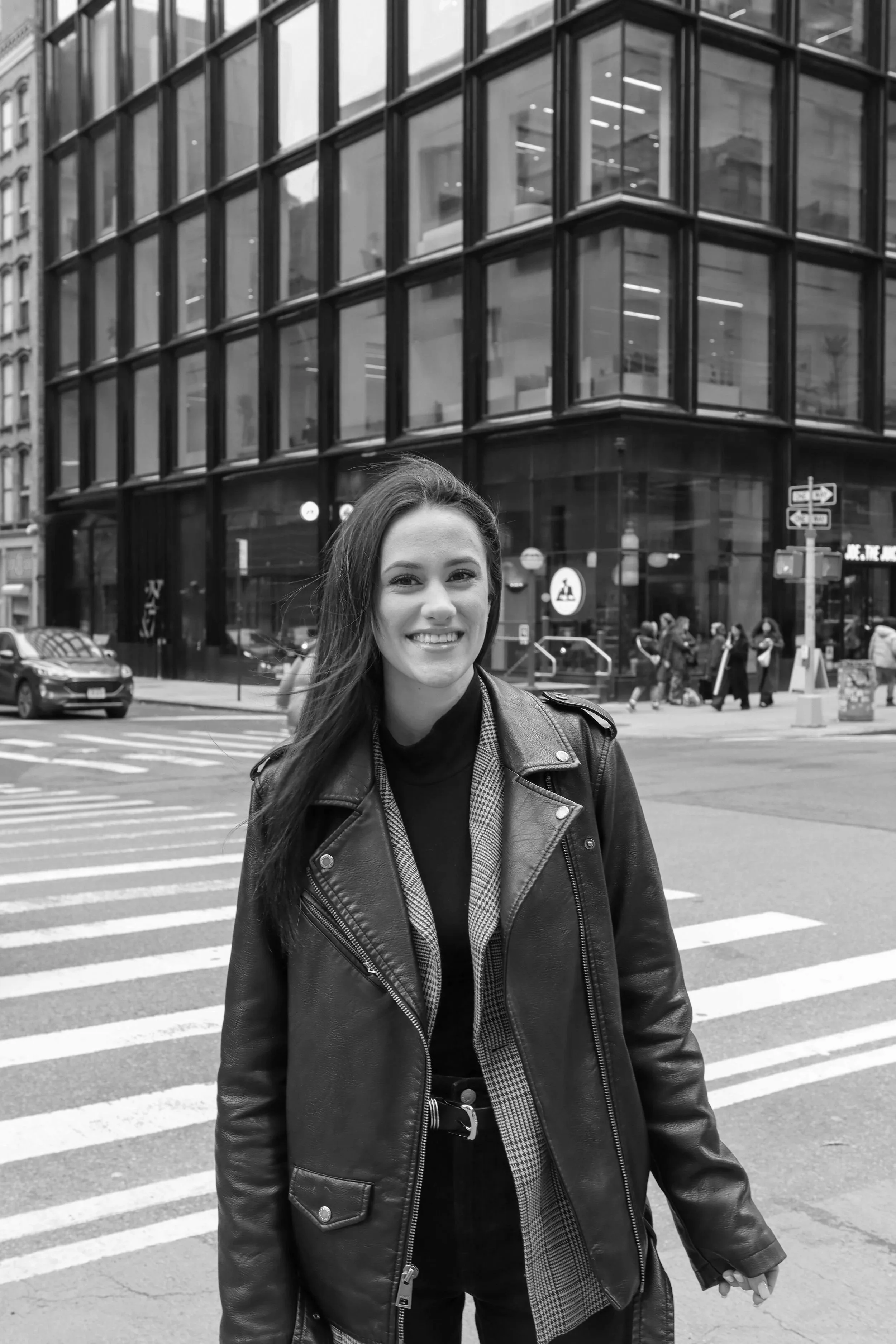 A woman (Katie Pasquarella) with long dark hair smiling, wearing a sequined top and a cross necklace, in a room with artwork in the background.