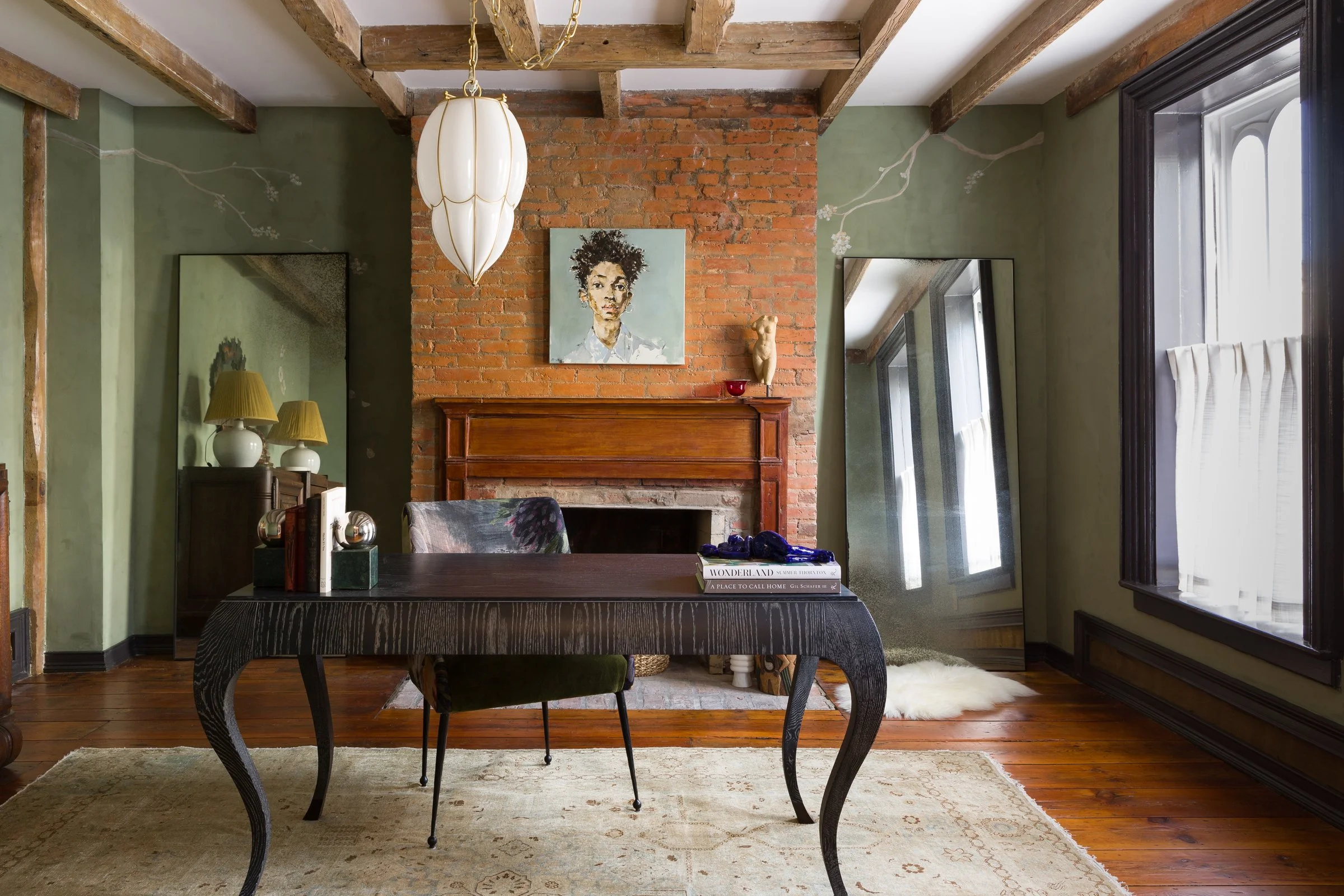 Interior of a rustic home office featuring a wooden desk, green upholstered chair, brick fireplace with a framed artwork, large mirror, and a window with curtains.