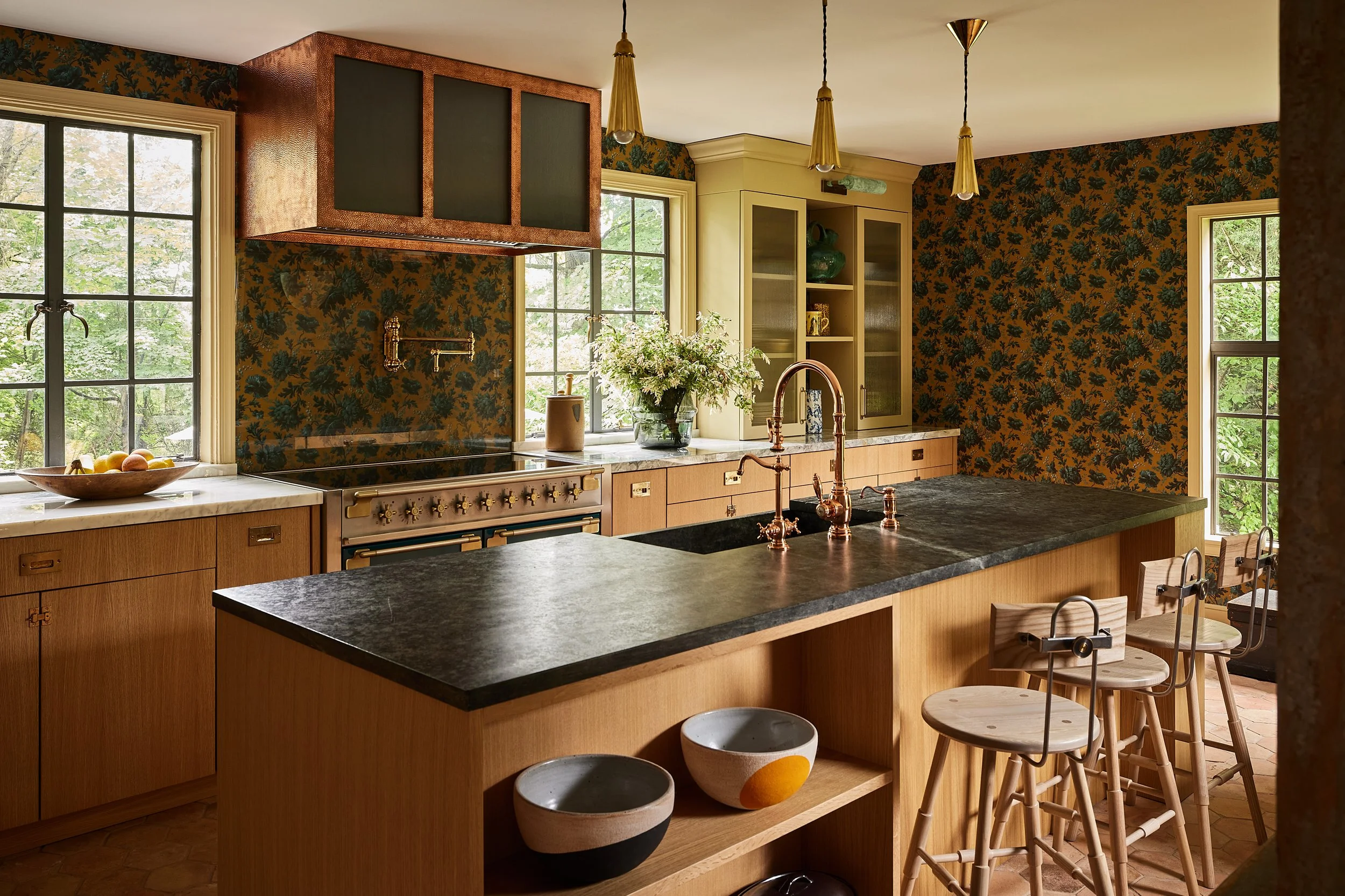 A kitchen with wooden cabinets, black countertops, a black stove, and a large island with a black surface and wooden base. There are three wooden bar stools at the island. The kitchen has large windows with black grid patterns, floral wallpaper with dark green leaves on a brown background, and hanging gold pendant lights.