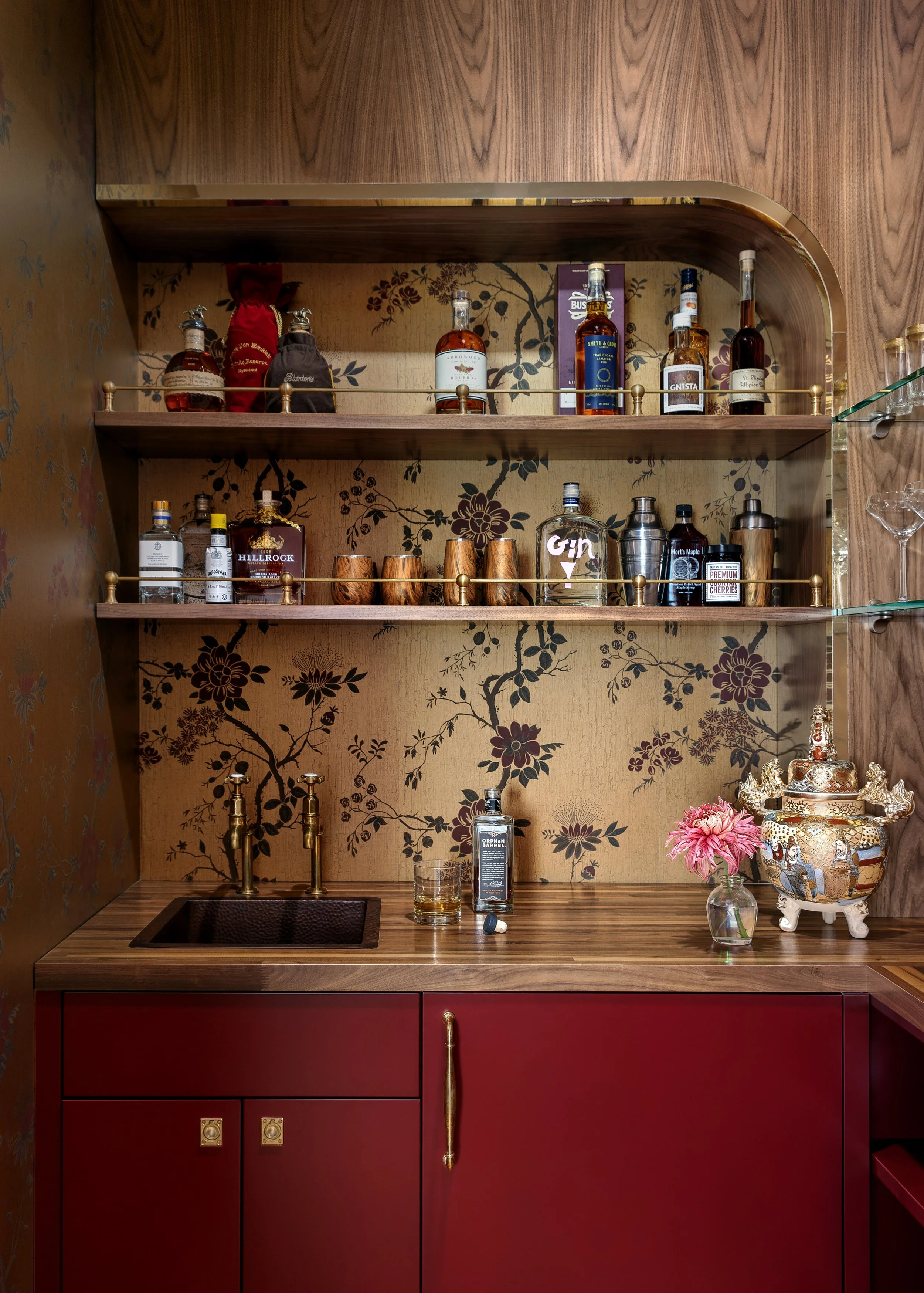 A stylish home bar with wooden shelving, a gold railing, and a red cabinet with gold handles. The shelves hold various liquor bottles, shot glasses, and cocktail tools. A small sink, a glass, a bottle of whiskey, a pink flower in a vase, and a decorative ceramic jar are on the countertop.