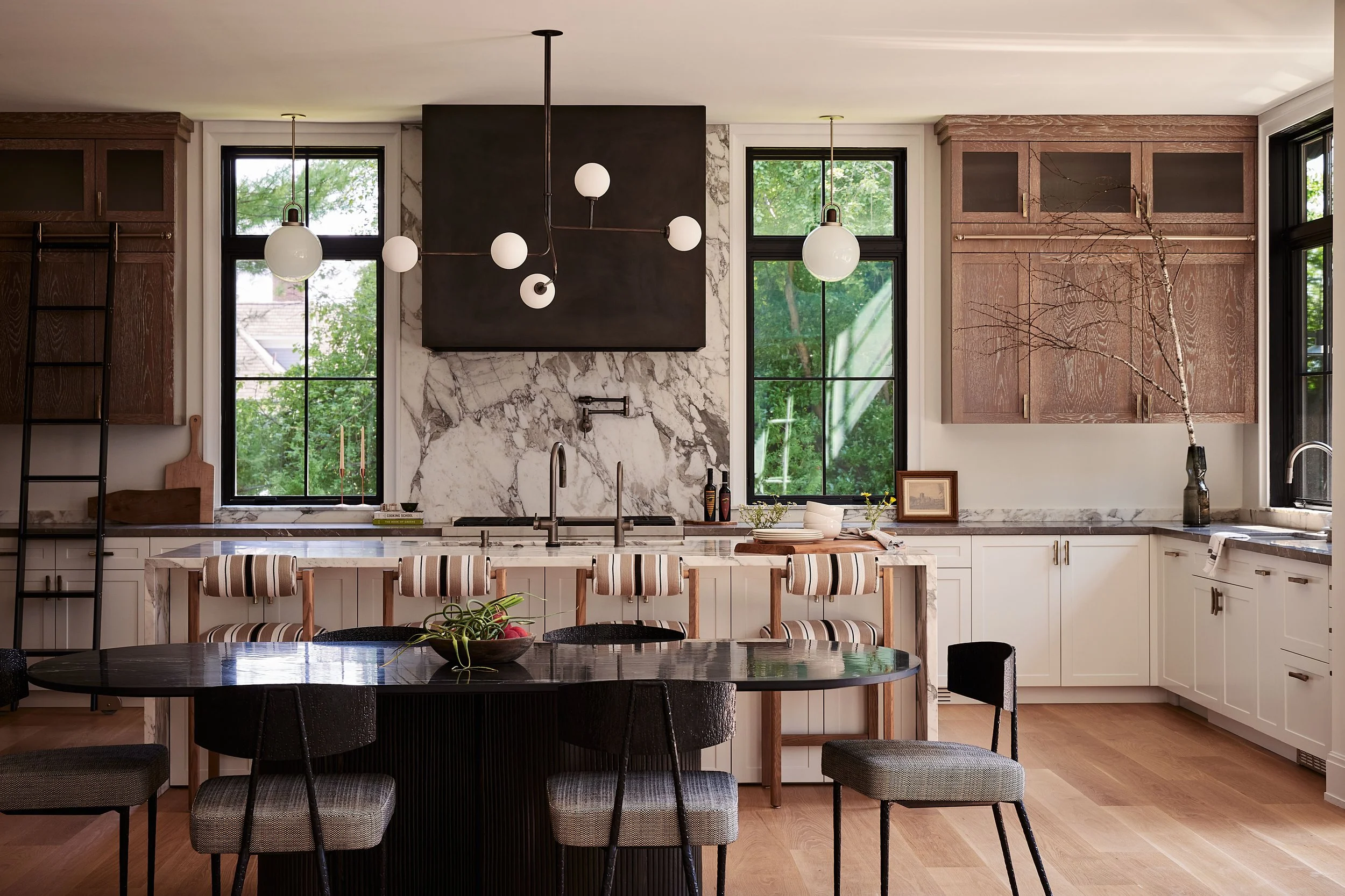 Modern kitchen with black and white marble backsplash, white lower cabinets, wooden upper cabinets, black dining table, and various chairs, with three large windows showing green trees outside.