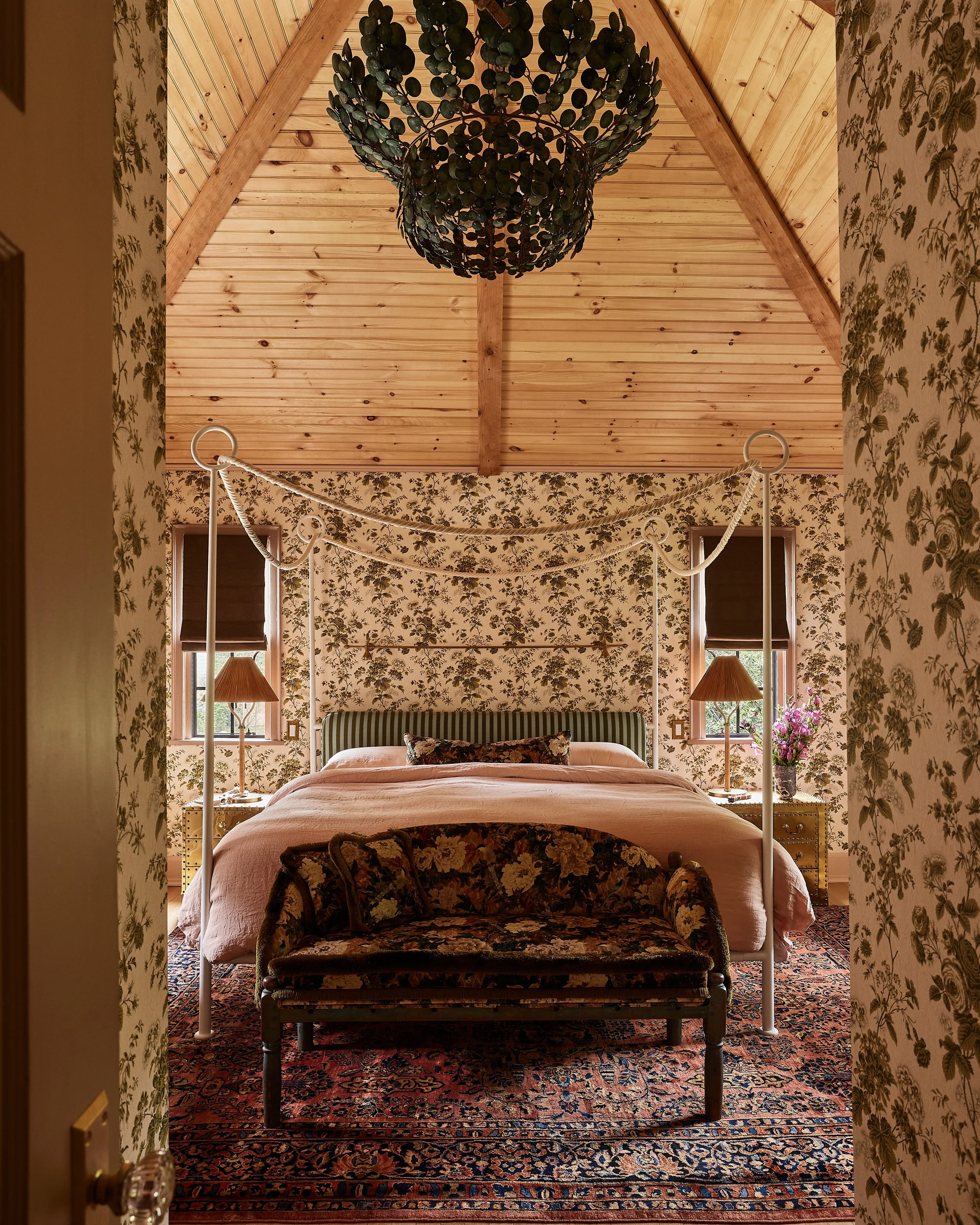 A cozy bedroom with floral wallpaper, a four-poster bed with a green headboard, a floral bench at the foot, two small nightstands with lamps, and two windows with brown blinds. The ceiling is wooden, and there's a dark green chandelier hanging from i