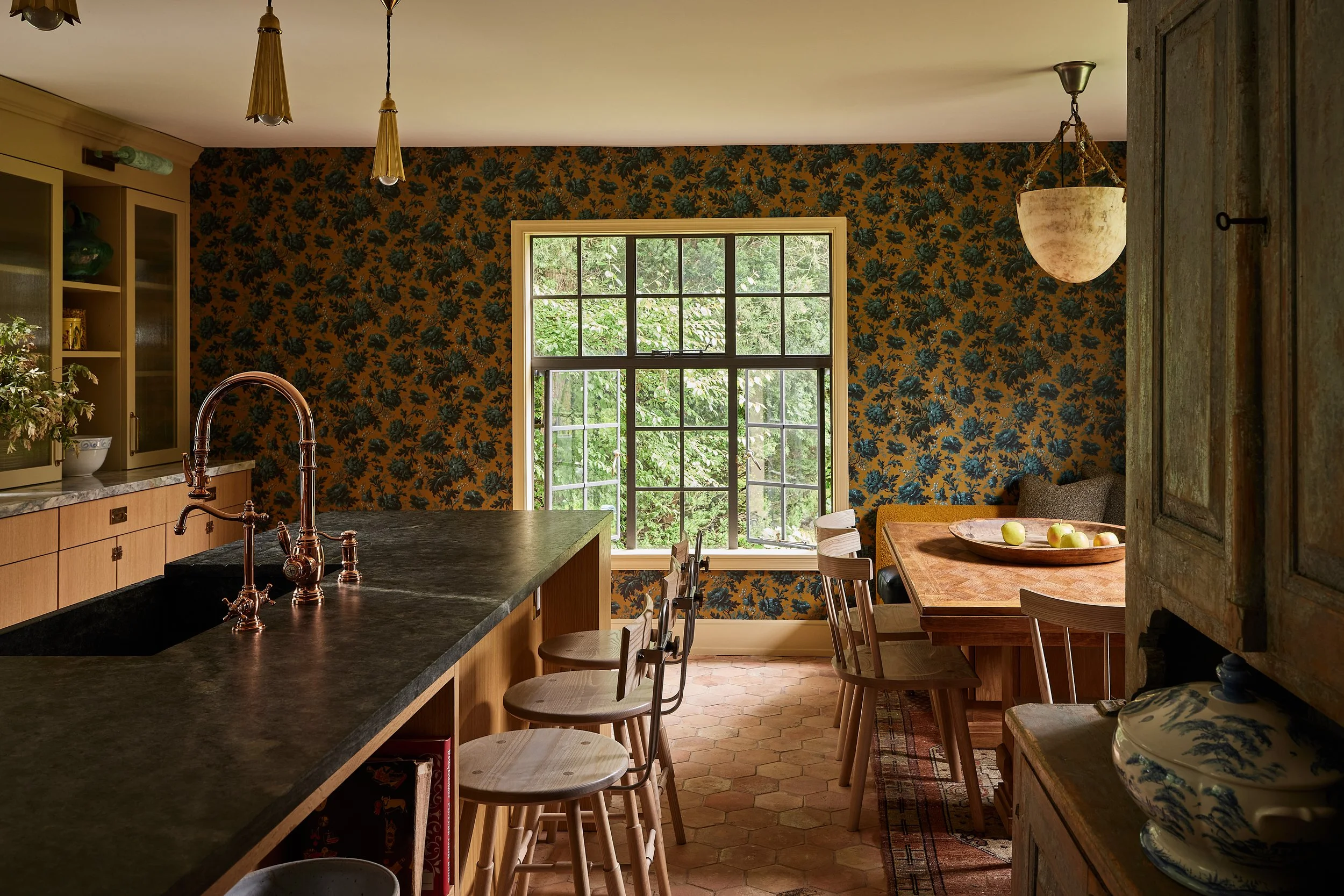 A kitchen with a dark stone countertop, a window with trees outside, floral wallpaper, a wooden dining table with chairs, and pendant lights hanging from the ceiling.