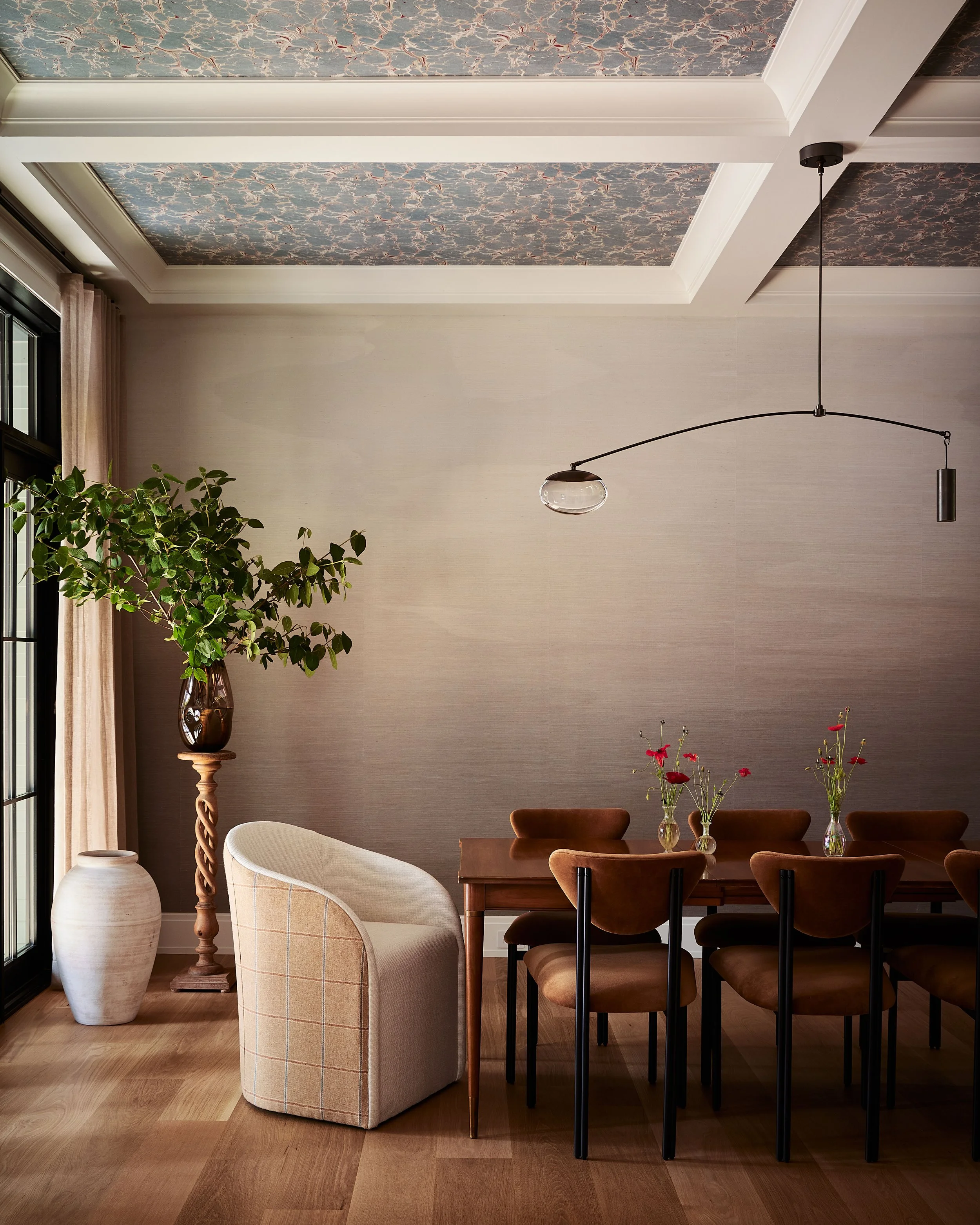 Living room with a wooden dining table, brown chairs, a cream armchair, large potted plant, decorative vases, and a modern ceiling light fixture.