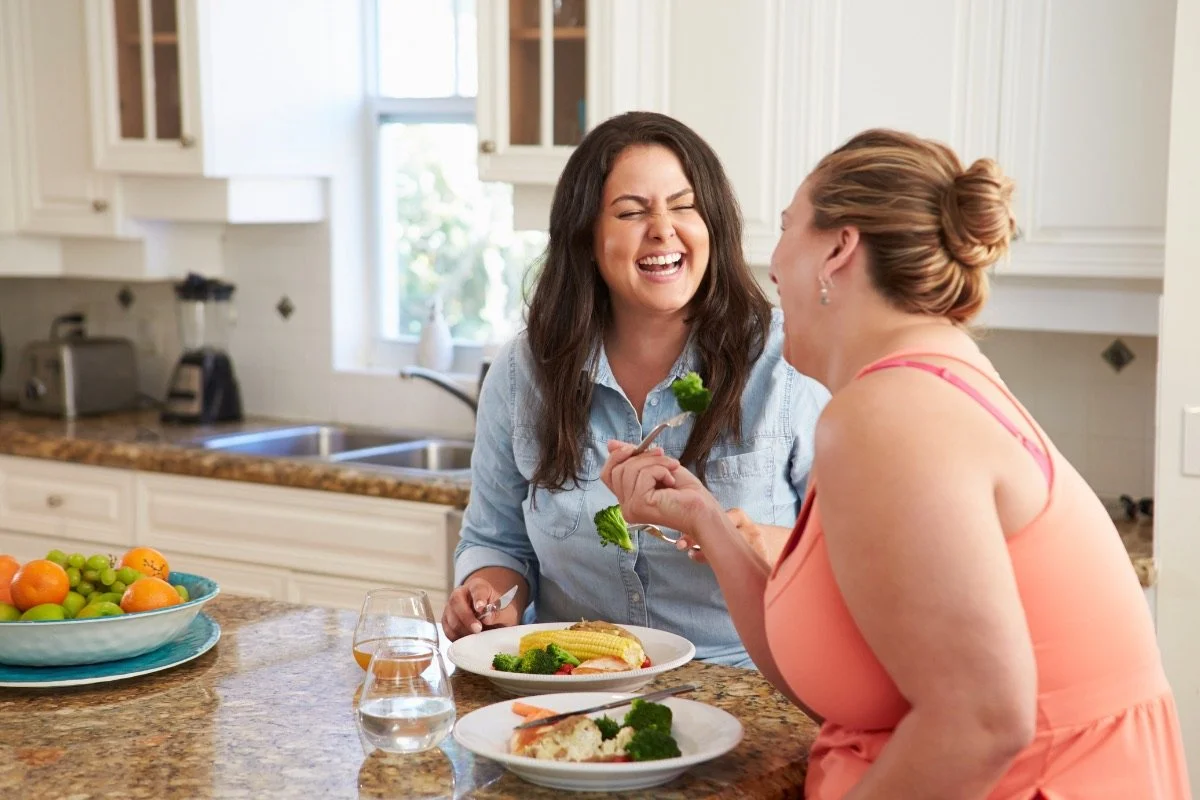 two women sitting at kitchen bench eating a healthy meal and laughing