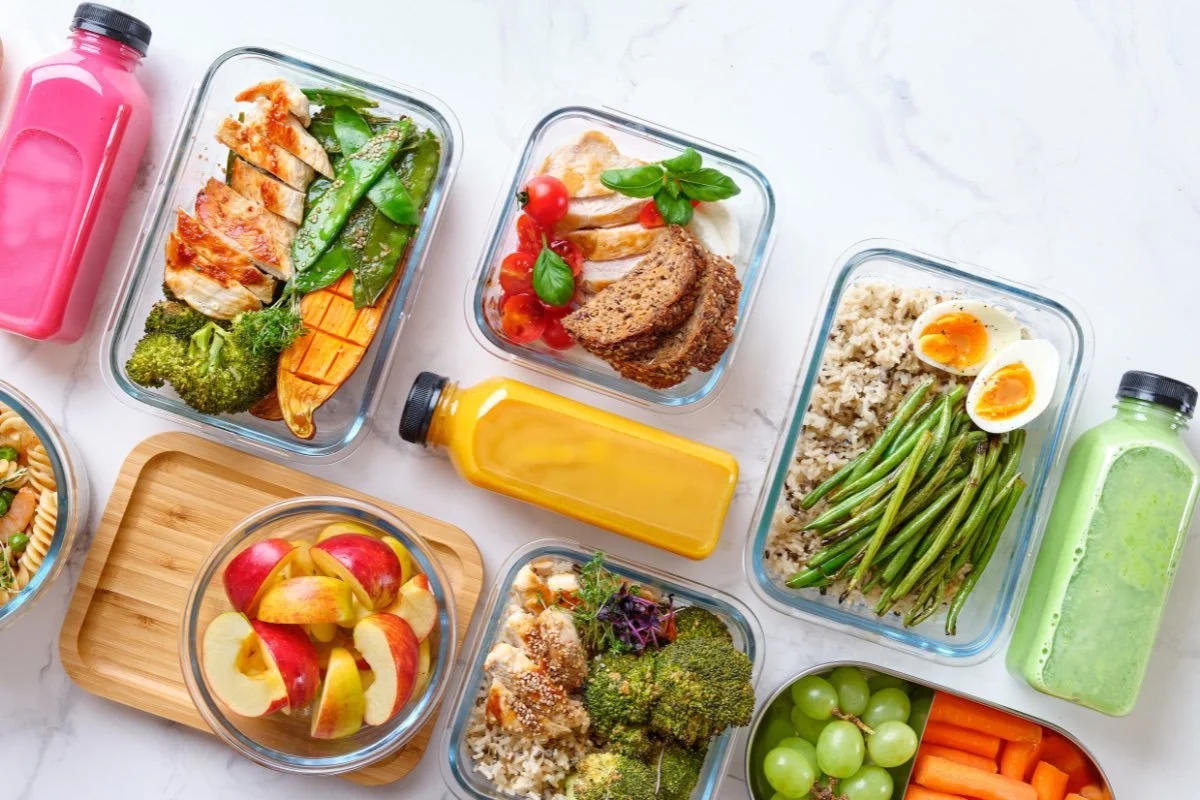 Assorted healthy meal prep containers on a white surface, including sliced fruits, cooked vegetables, boiled eggs, and various protein dishes with bottles of juice. An example of a Wegovy diet plan