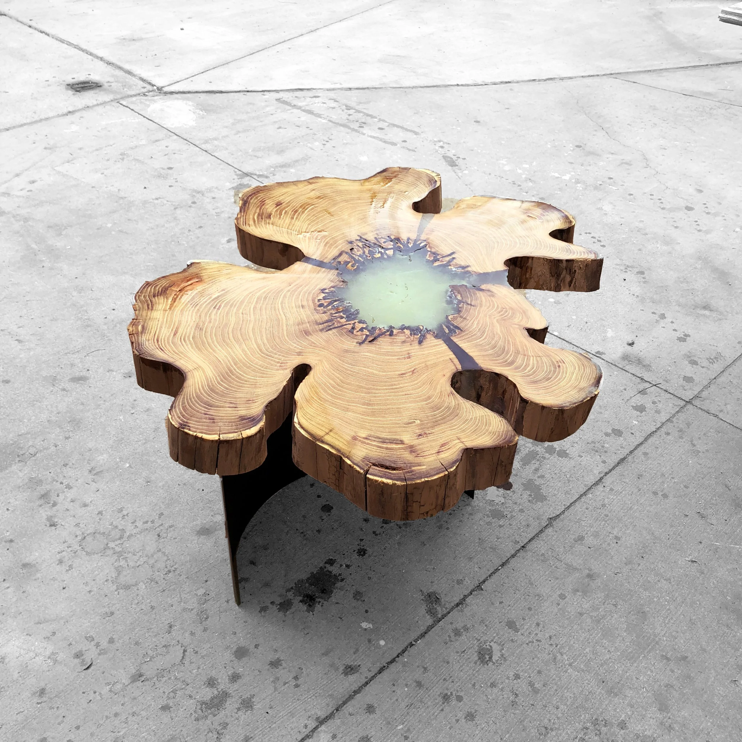 A wooden table carved in the shape of a flower with visible tree rings and a resin-filled center, situated on a concrete surface.