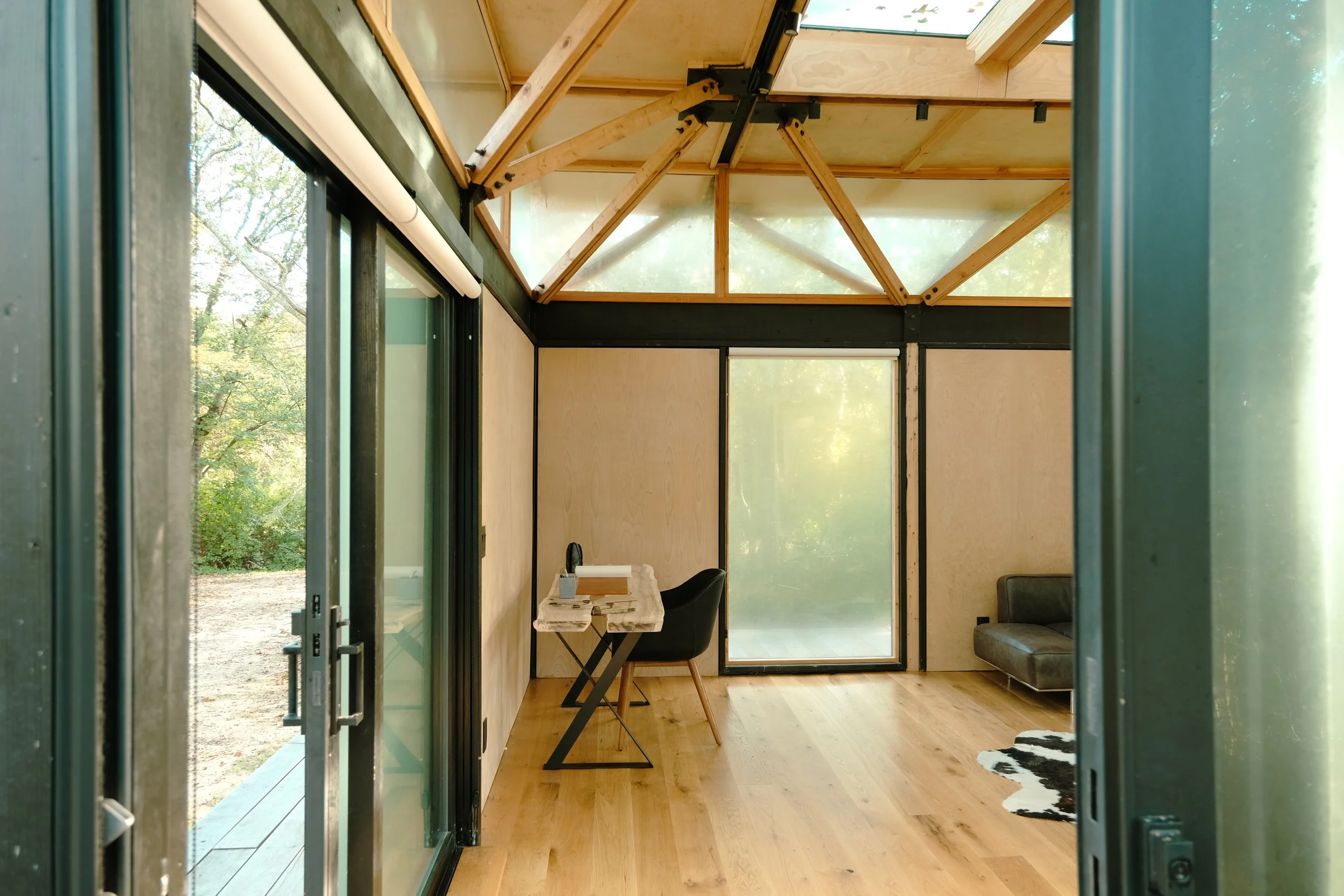 Interior of a modern, minimalist room with large glass sliding doors, wooden floors, and light-colored walls. There is a small black desk with a chair, a black sofa, and a black and white cowprint rug. The ceiling has exposed wooden beams and trusses, with a large skylight letting in natural light.