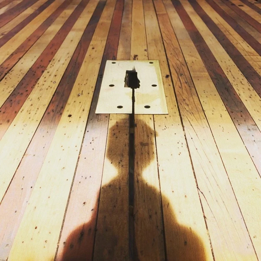 Close-up of a wooden floor with a rectangular cutout and an electrical cable running through it, shadows of two people cast on the floor.
