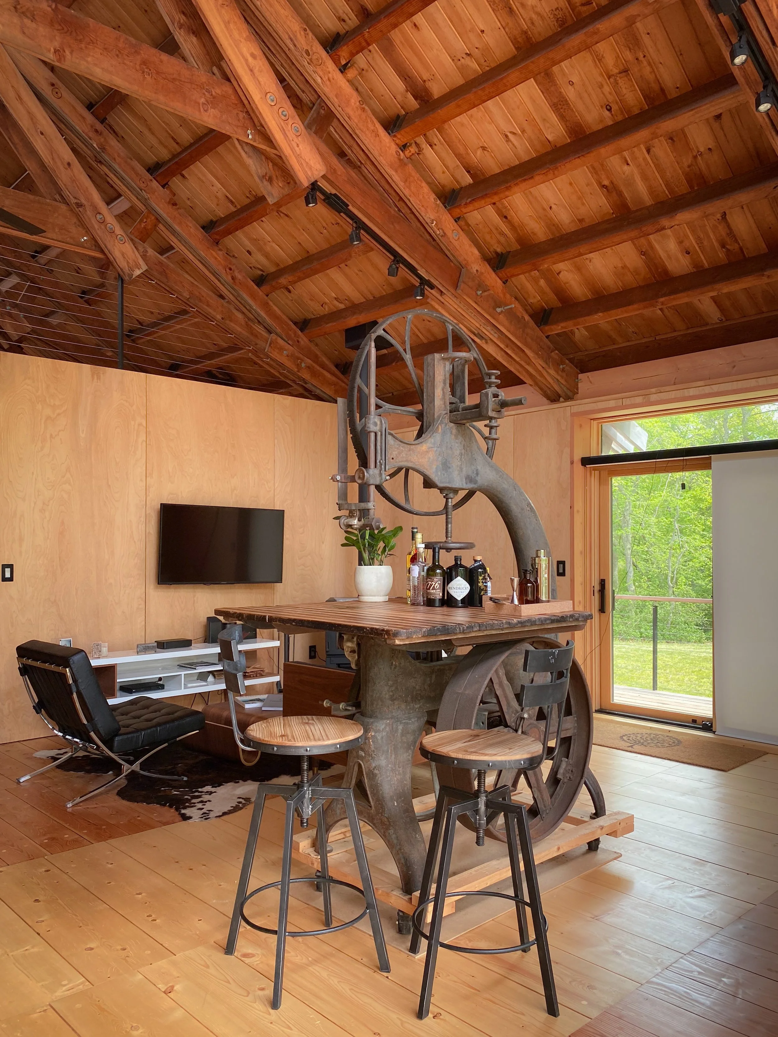 Interior of a room with wooden walls and ceiling, featuring an antique meat slicer turned into a bar, with bottles on top, and three tall stools around it, near a sliding glass door with a view of green trees outside.