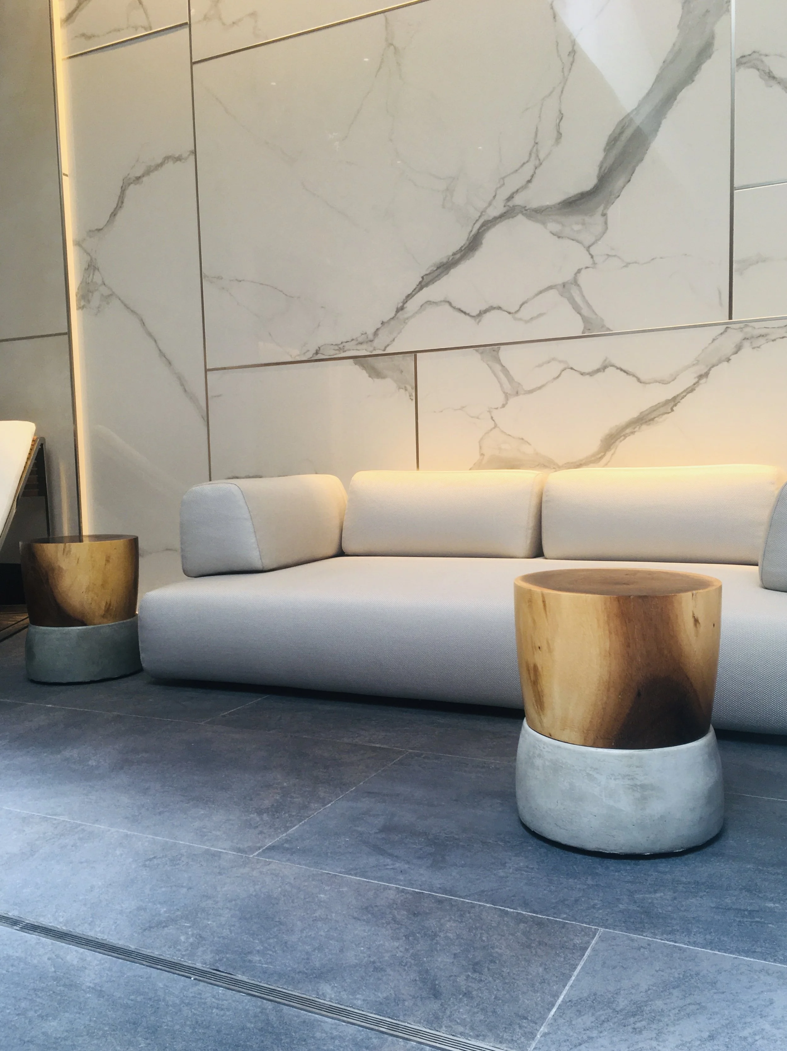 Modern lobby with beige sofa, two wooden and concrete side tables, and marble wall tiles.