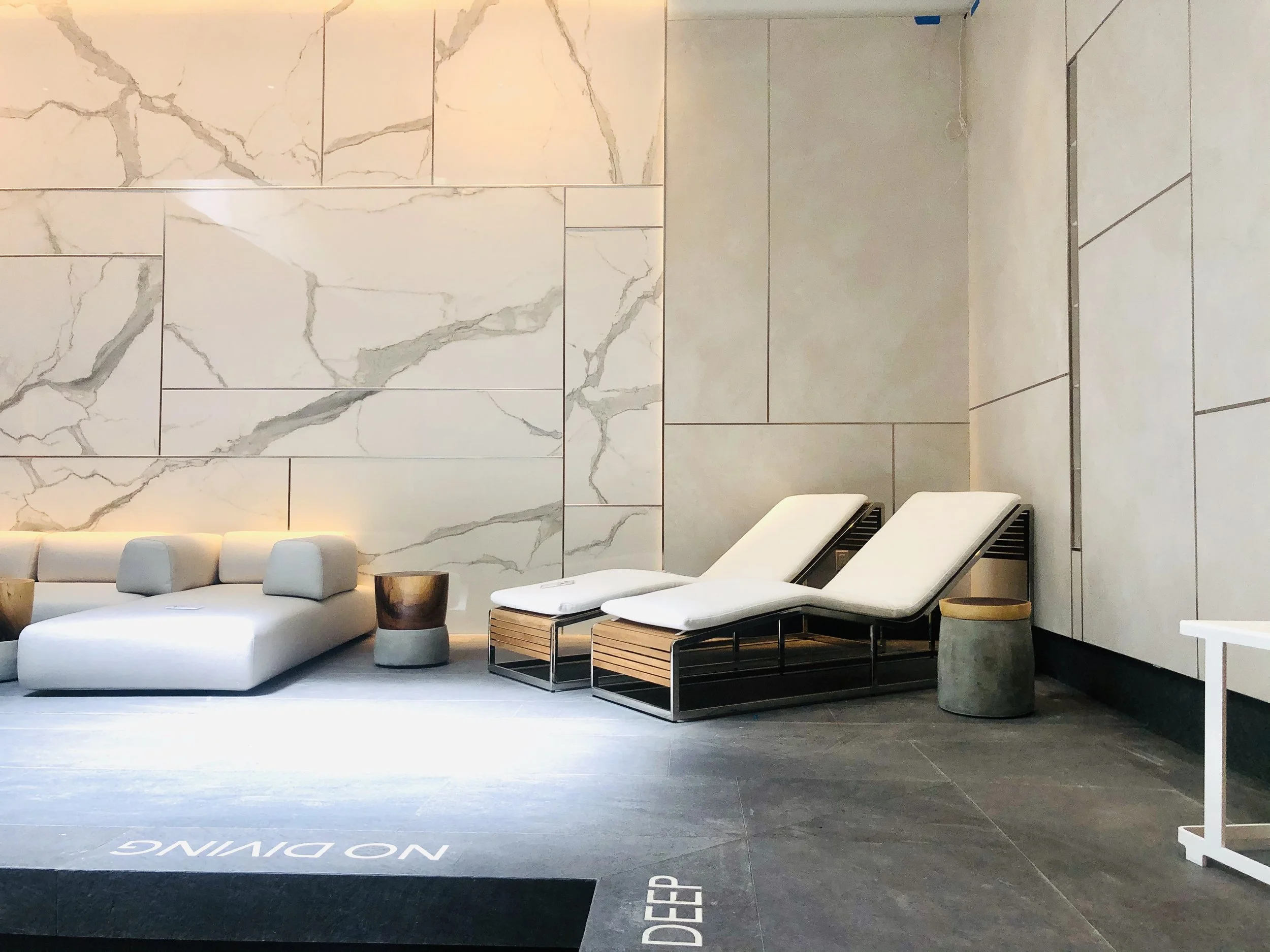 Lobby with modern furniture, including white lounge chairs, a sofa with cushions, and small side tables, set against a marble wall and dark flooring.