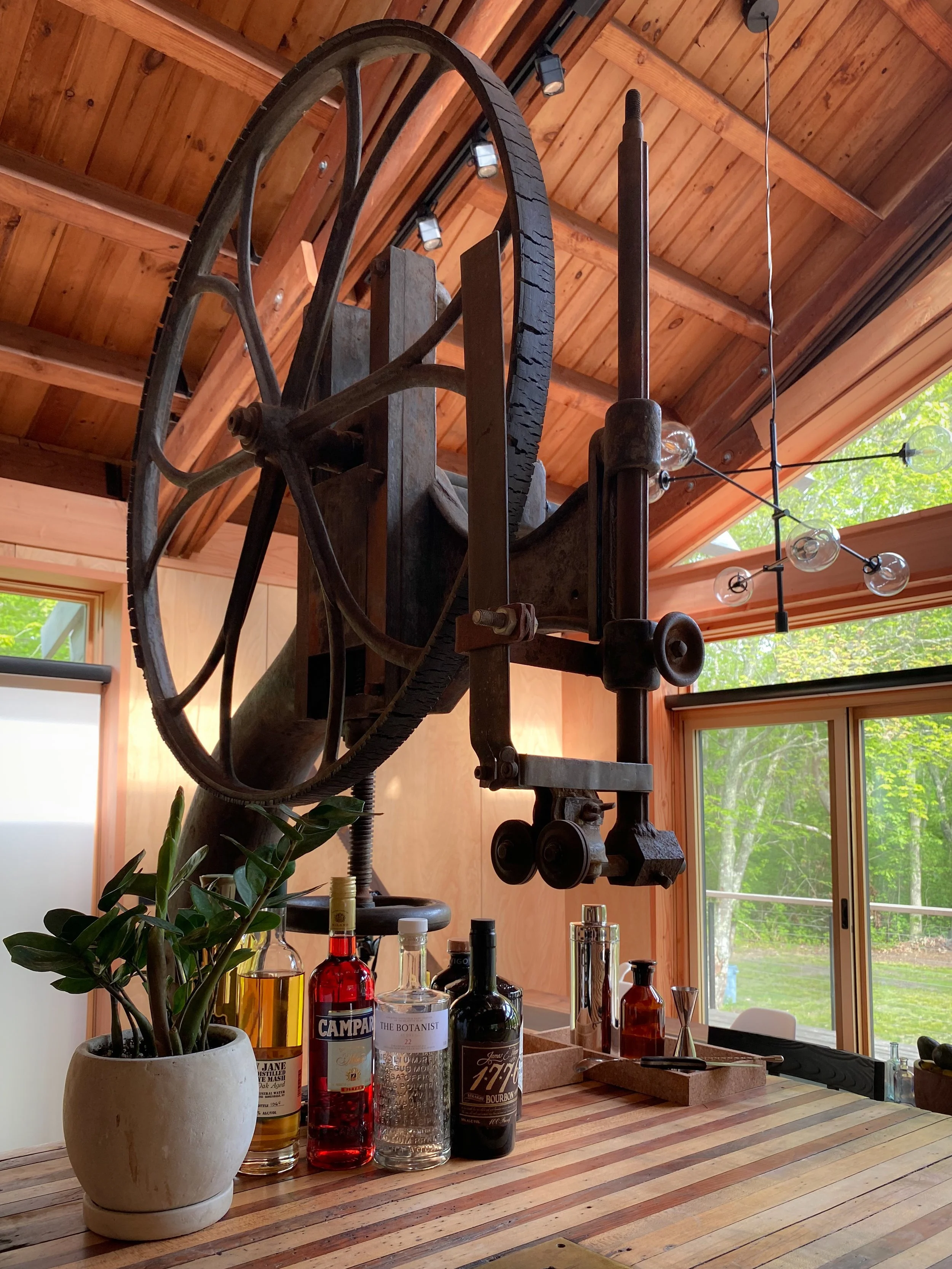 A rustic wooden room with a vintage mechanical device hanging from the ceiling above a kitchen counter. On the counter are several bottles of alcohol, a potted plant, and bar tools, with large windows showing green trees outside.