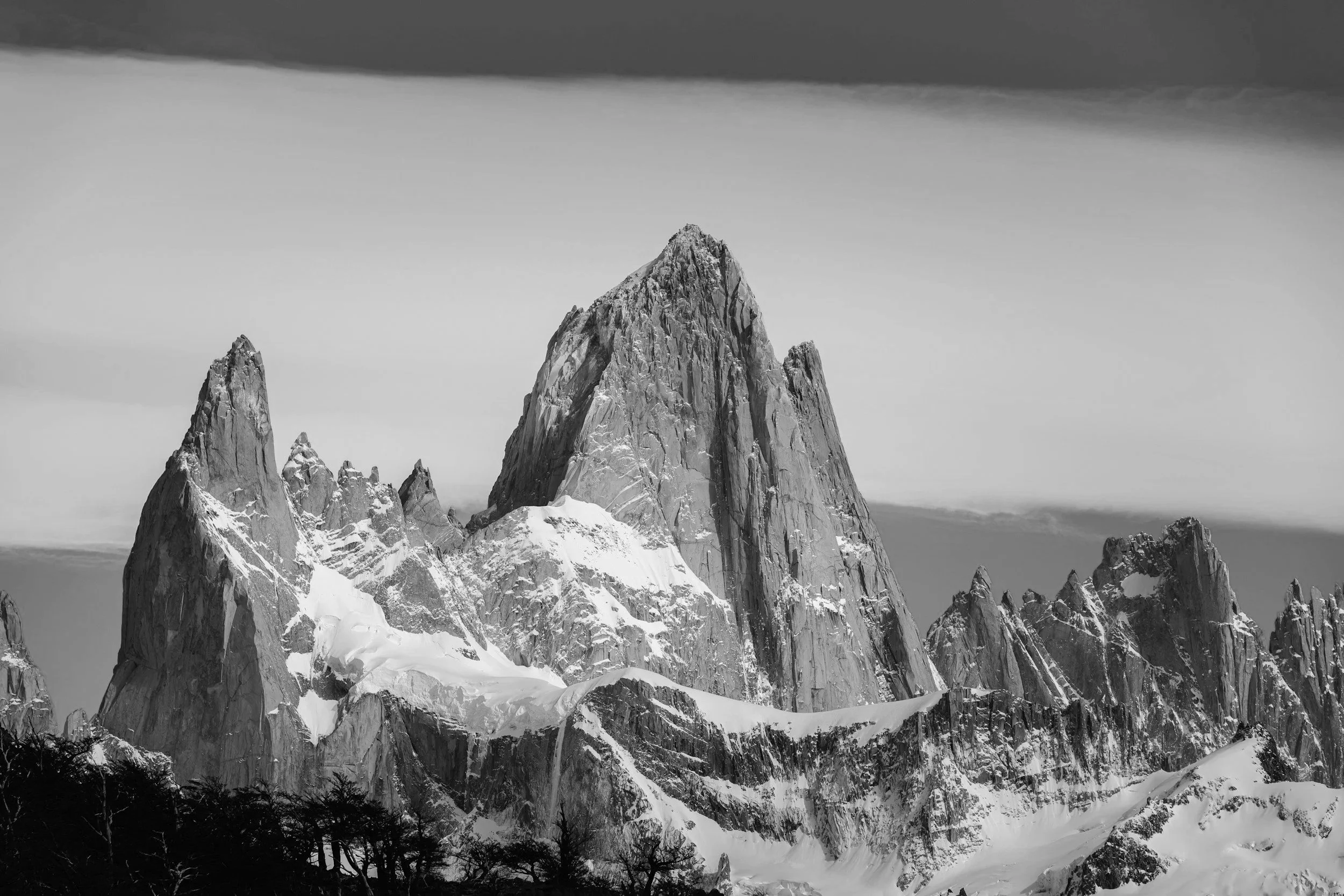 Black and white photograph of rugged snow-covered mountain peaks, with a cloudy sky in the background.