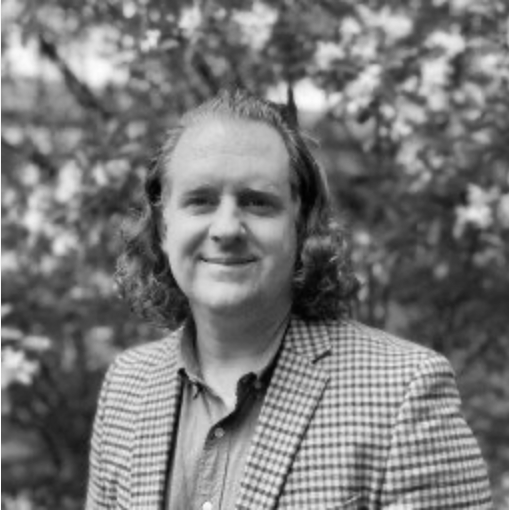A man with shoulder-length hair and a checkered blazer standing outdoors with trees in the background.