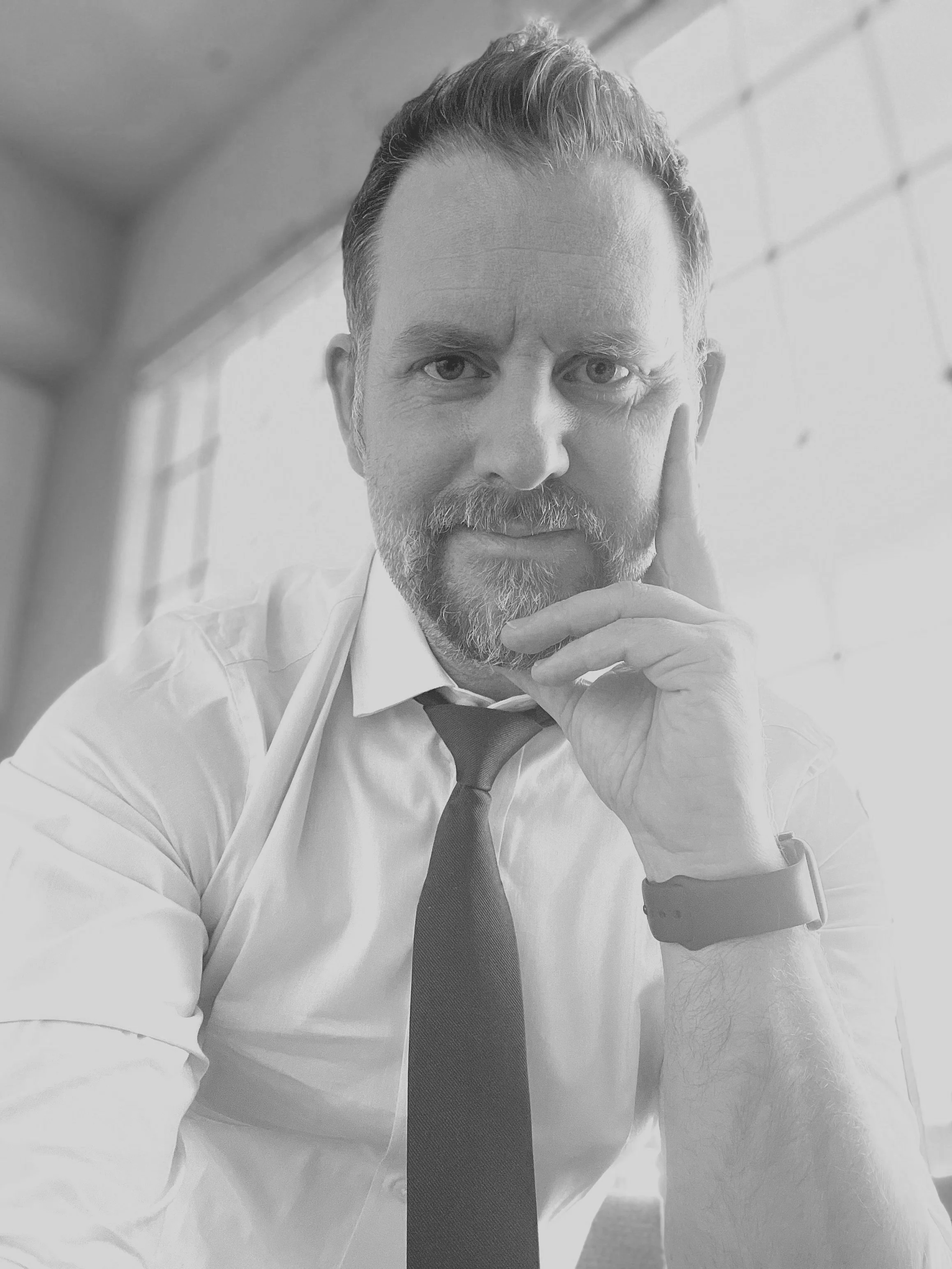 A black-and-white photo of a man with a beard and mustache, wearing a white shirt, a dark tie, and a smartwatch, sitting near a window and looking at the camera.