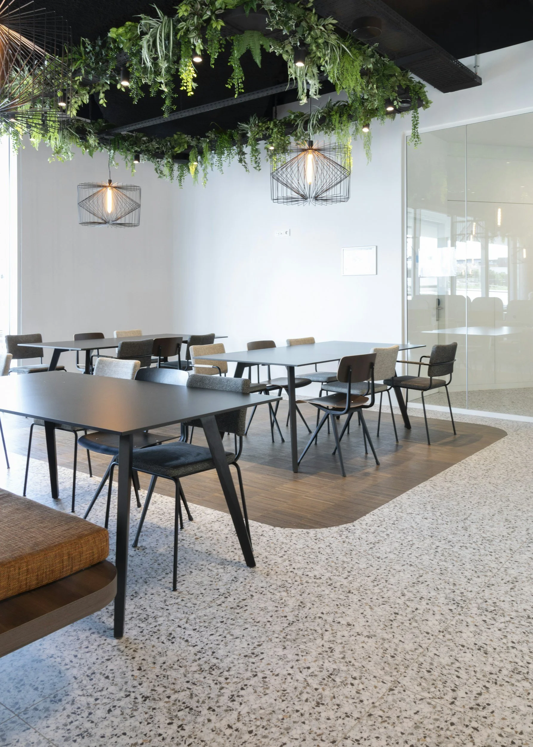 Modern meeting room with black tables, a variety of chairs, hanging industrial-style light fixtures, and greenery hanging from the ceiling.