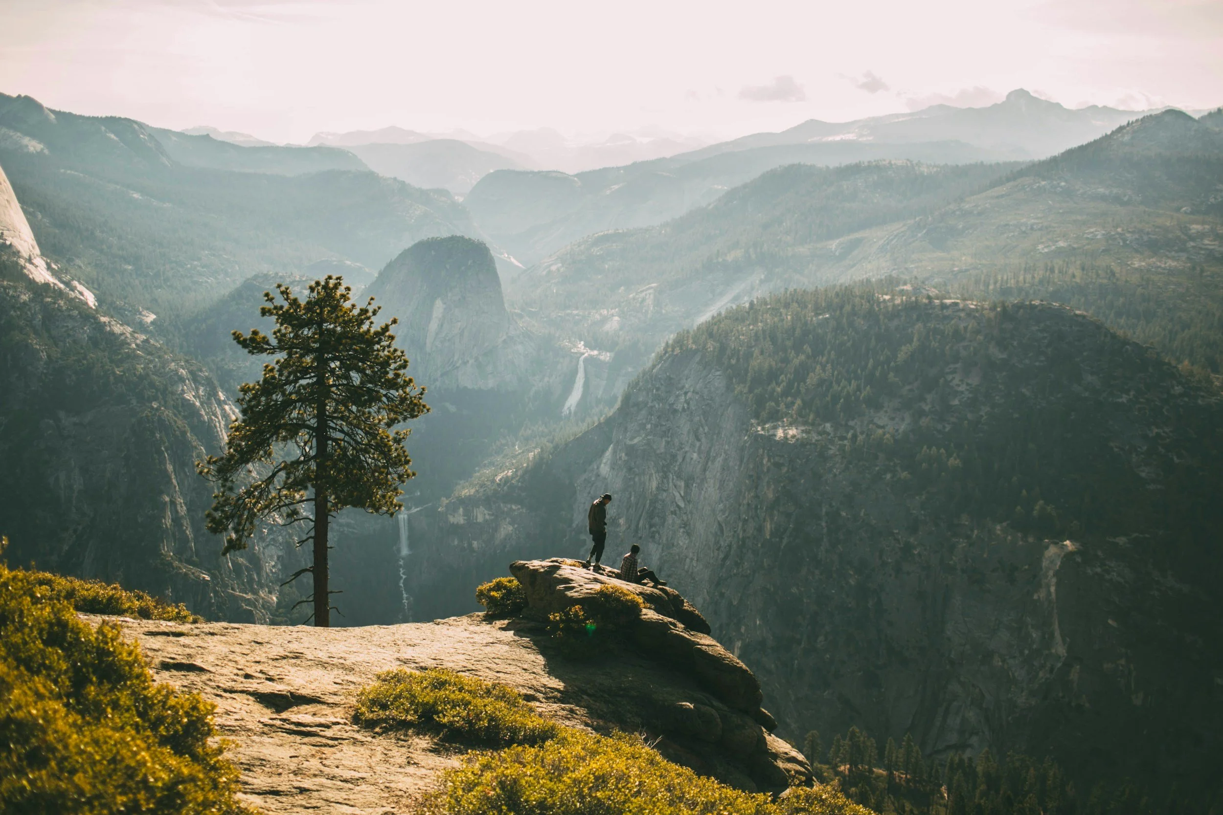 Two people sitting on a rocky ledge overlooking a deep canyon with waterfalls and mountainous forested landscape in the background, during daylight.