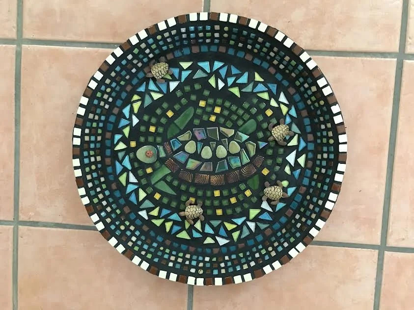 Mosaic wall clock with a colorful geometric pattern, featuring four turtle figurines at the 12, 3, 6, and 9 o'clock positions, on a tiled floor.