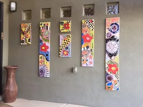 A wall displaying seven colorful abstract paintings arranged in descending order of height, with a tall, brown textured vase on the left side and a small, rectangular wall fixture above.