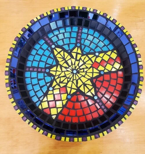 Colorful mosaic clock with a yellow star at the center, surrounded by blue, red, and yellow sections, all made from small tiles.