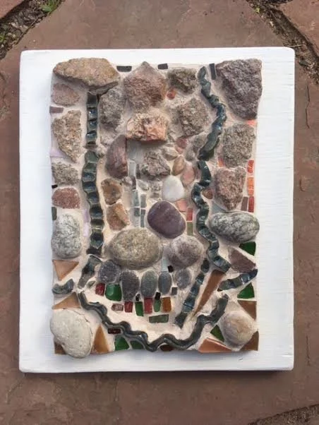 A mosaic artwork made of various stones and pebbles arranged on a white square surface with colorful glass pieces underneath.