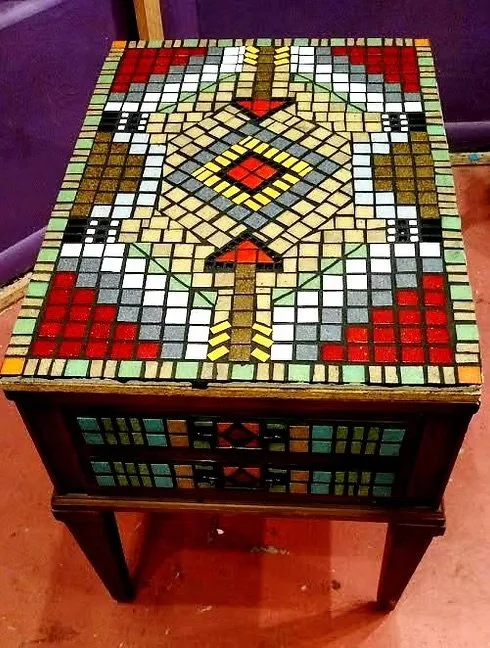 A wooden table with a mosaic tile top featuring a symmetrical geometric pattern in red, black, white, yellow, and green colors.