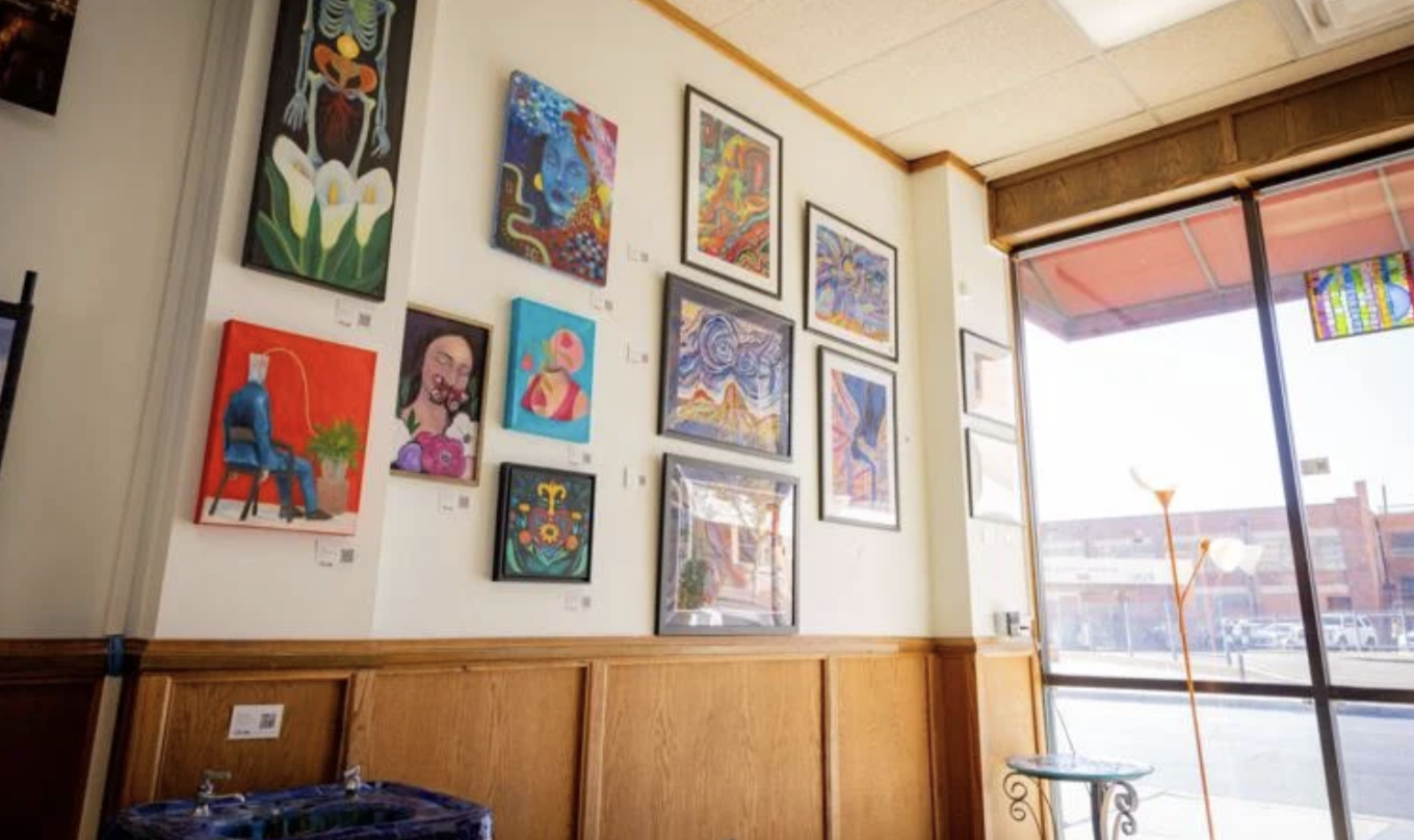 An art gallery with colorful paintings on the wall, large windows allowing outside light, and a modern floor lamp inside.