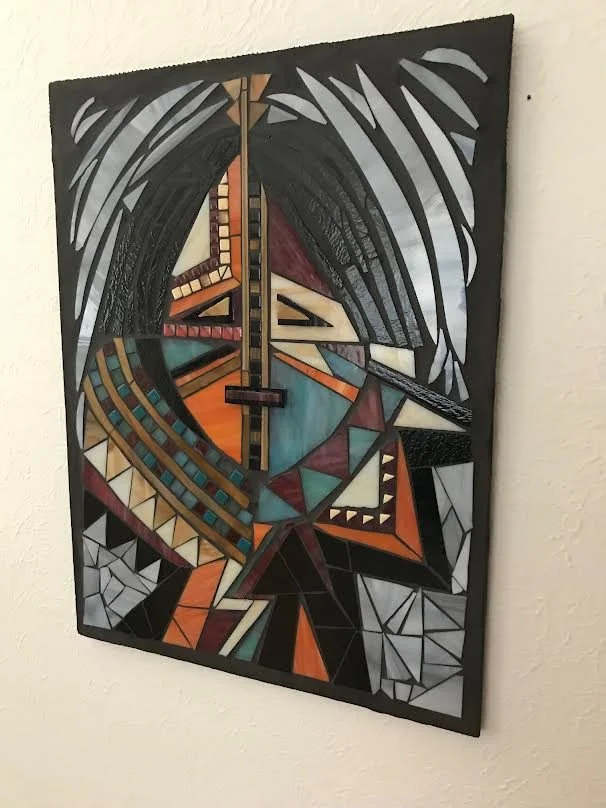 A geometric abstract collage artwork featuring sharp lines, triangles, and various colors including black, white, orange, and teal, mounted on a wall.