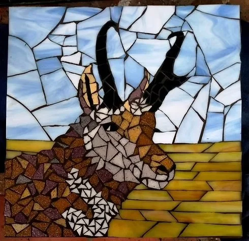 Mosaic artwork of a goat with black horns standing on a field with a blue sky background.