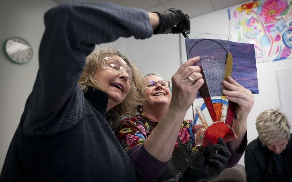 Two women are smiling and holding up a colorful stained glass artwork while a third woman is seated nearby, looking down. The setting appears to be an art studio with colorful paintings on the wall.