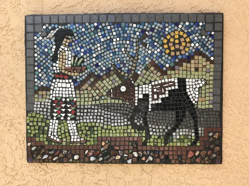 A mosaic depicting a person with long dark hair wearing a white shirt and patterned pants, standing next to a black donkey, against a background of mountains, blue sky, and the sun.