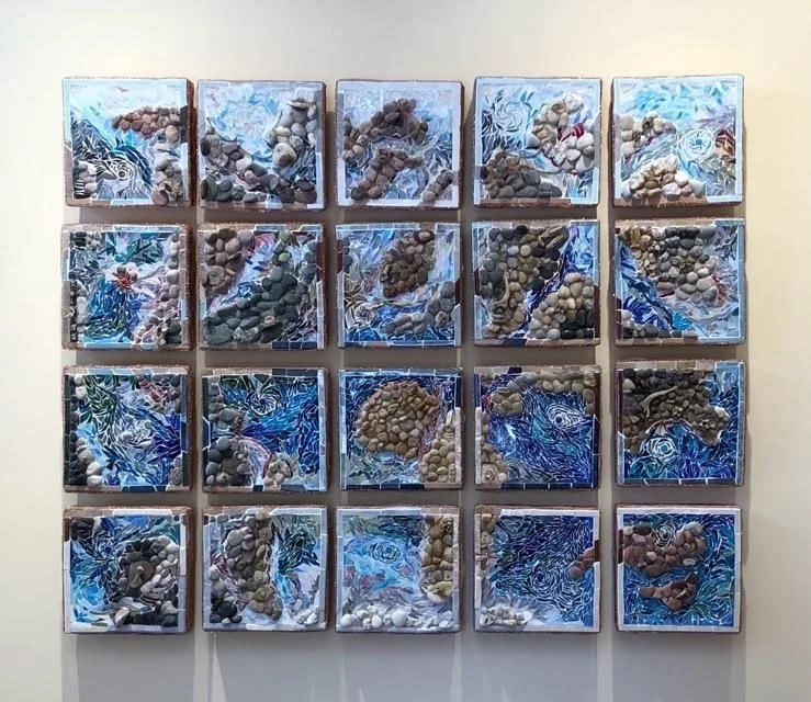 A wall display with 20 framed pieces arranged in four rows and five columns, each containing a three-dimensional collage of stones or pebbles on a blue, white, and black patterned background.