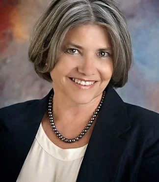 Portrait of a smiling woman with short gray hair wearing a dark blazer and a pearl necklace, against a blurred background.